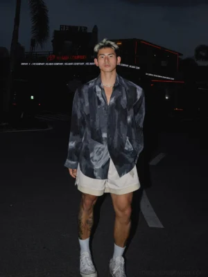 Male model wearing an abstract printed oversized satin shirt with dark grey watercolor pattern, beige shorts, white socks and sneakers on a nighttime street