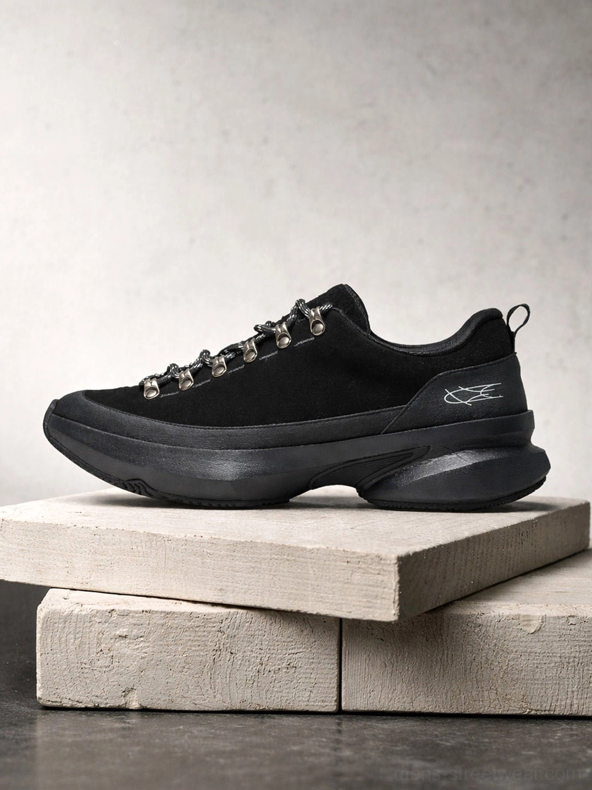 All-black casual hiking-inspired streetwear sneaker displayed on concrete blocks with matte upper, metal lace hardware, textured details, and chunky outsole