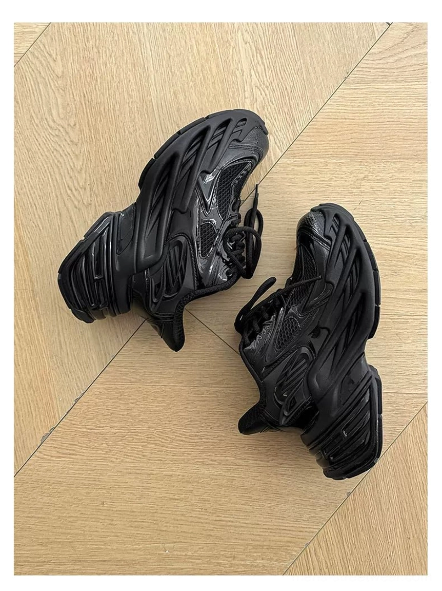 All-black futuristic chunky runner sneakers with angular upper and oversized platform outsole on wood flooring