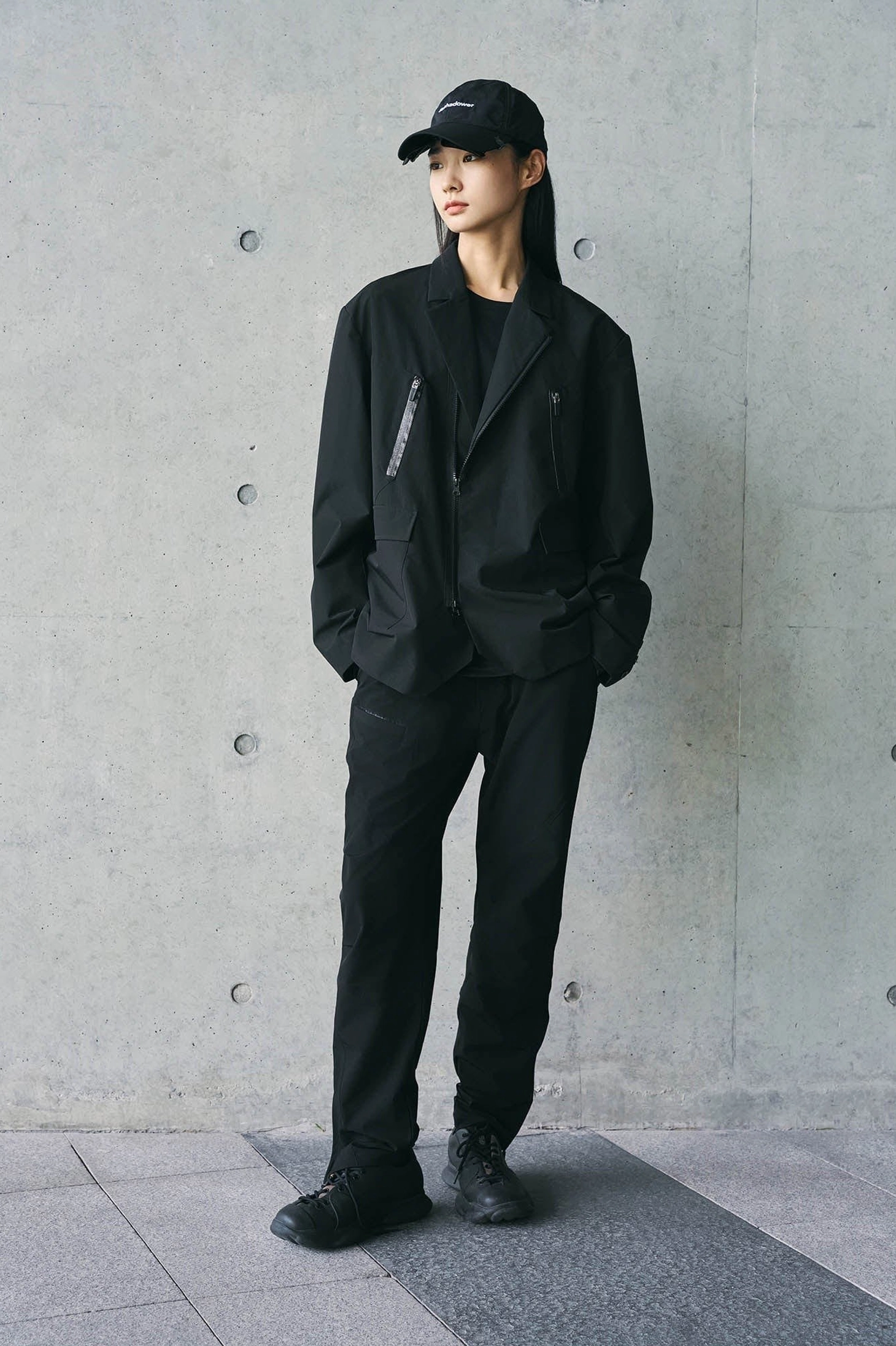 Full body view of an all-black Korean streetwear outfit with technical track pants oversized jacket and baseball cap
