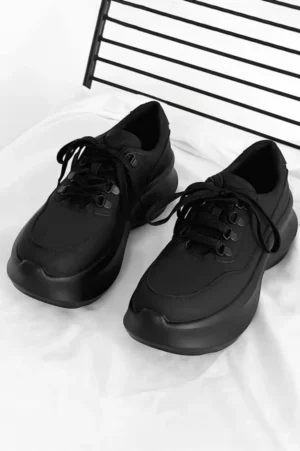 All-black platform lace-up sneakers shown from a top-down view on a white fabric background, featuring a matte cowhide leather upper and chunky rubber sole