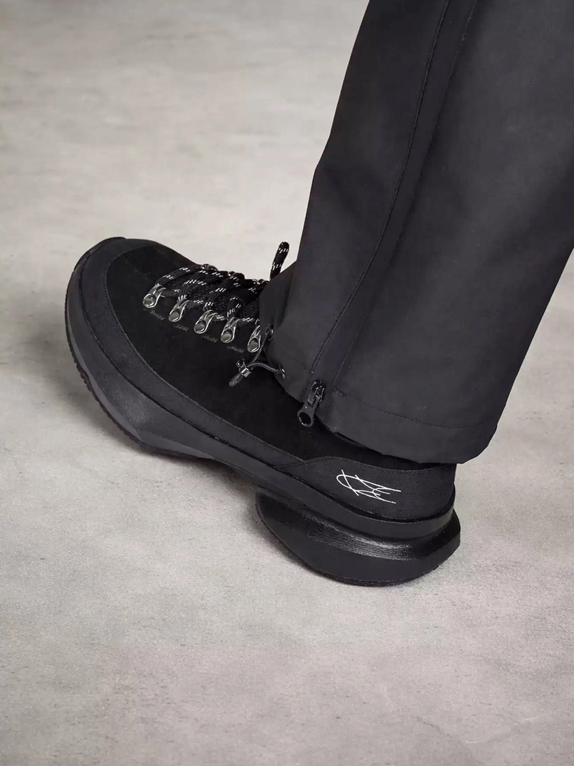 Rear side view of all-black streetwear sneakers styled with cropped tapered pants, highlighting the structured heel and thick curved rubber-style outsole