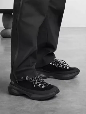 All-black techwear streetwear sneakers worn with tapered technical pants in a low-angle front three-quarter view, featuring metal lace hardware and a thick sculpted outsole