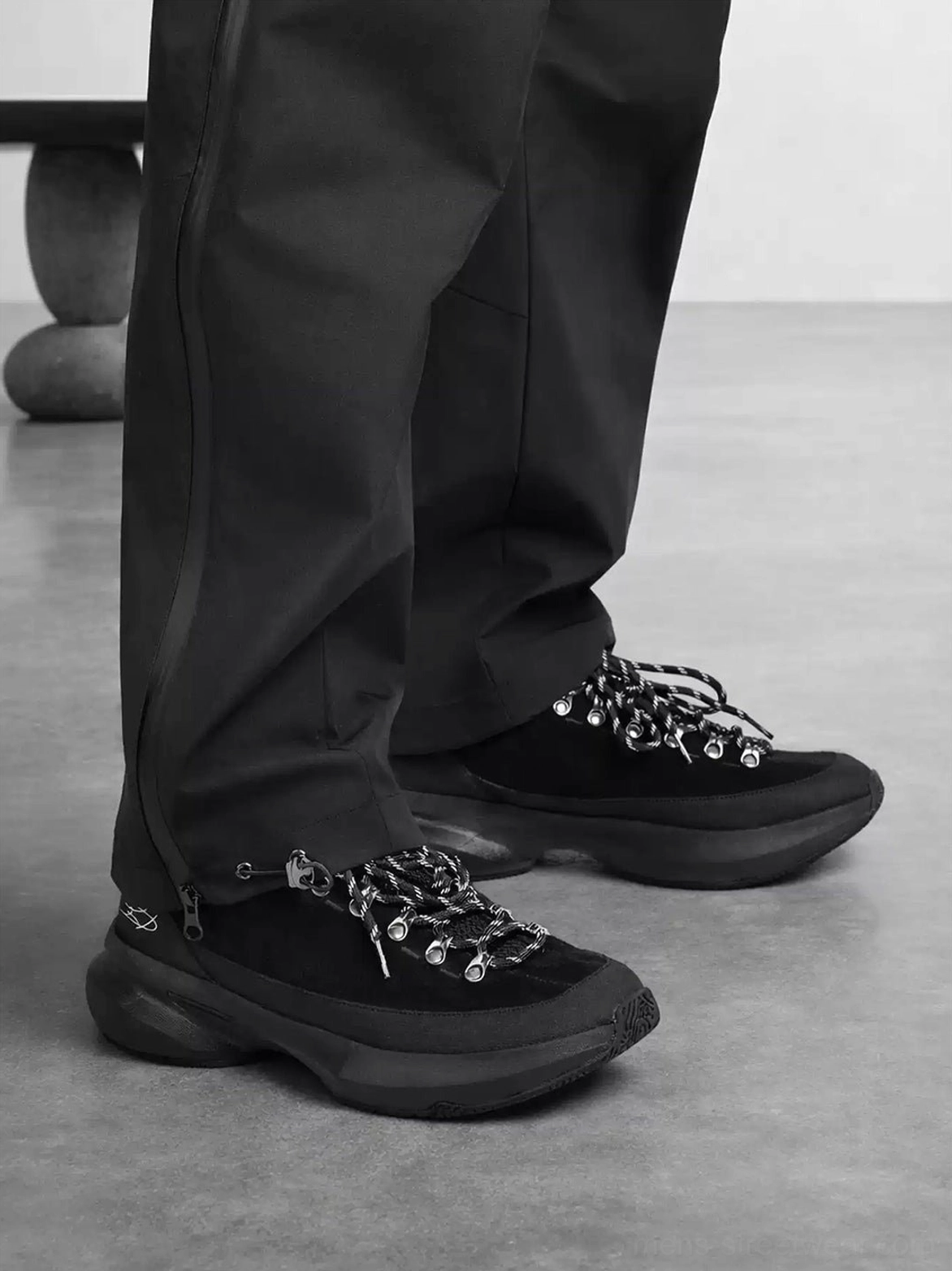 All-black techwear streetwear sneakers worn with tapered technical pants in a low-angle front three-quarter view, featuring metal lace hardware and a thick sculpted outsole
