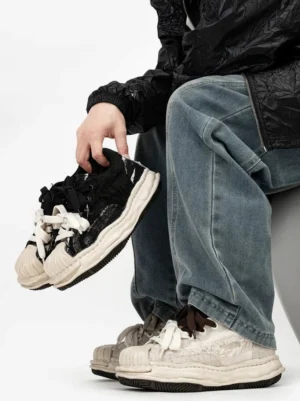 Distressed double-lace shell-toe chunky sneakers shown from a low angle with frayed upper, thick molded rubber sole, and layered black and cream laces