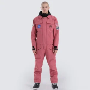 Dusty rose astronaut mission one-piece ski suit worn by male model, showing embroidered space patches, black hood lining, ribbed cuffs, and relaxed snow streetwear fit