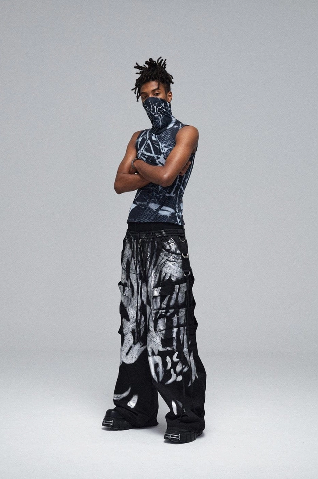Three-quarter front view of avant-garde black graffiti cargo pants with full silver artwork, wide-leg fit, and layered streetwear styling