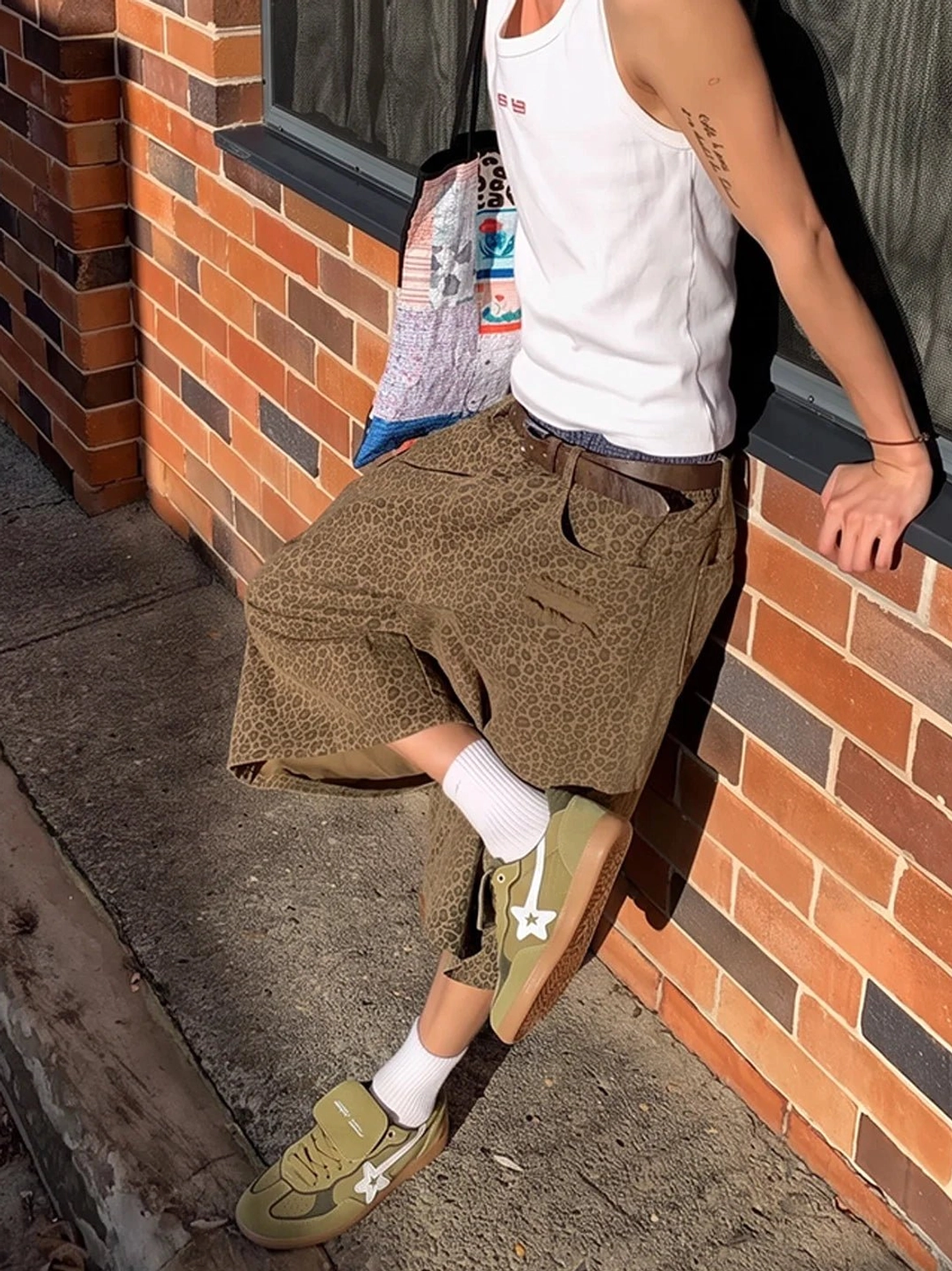 Male model wearing avocado suede star-panel retro sneakers with white socks, cropped brown trousers, and a white tank top on an outdoor sidewalk