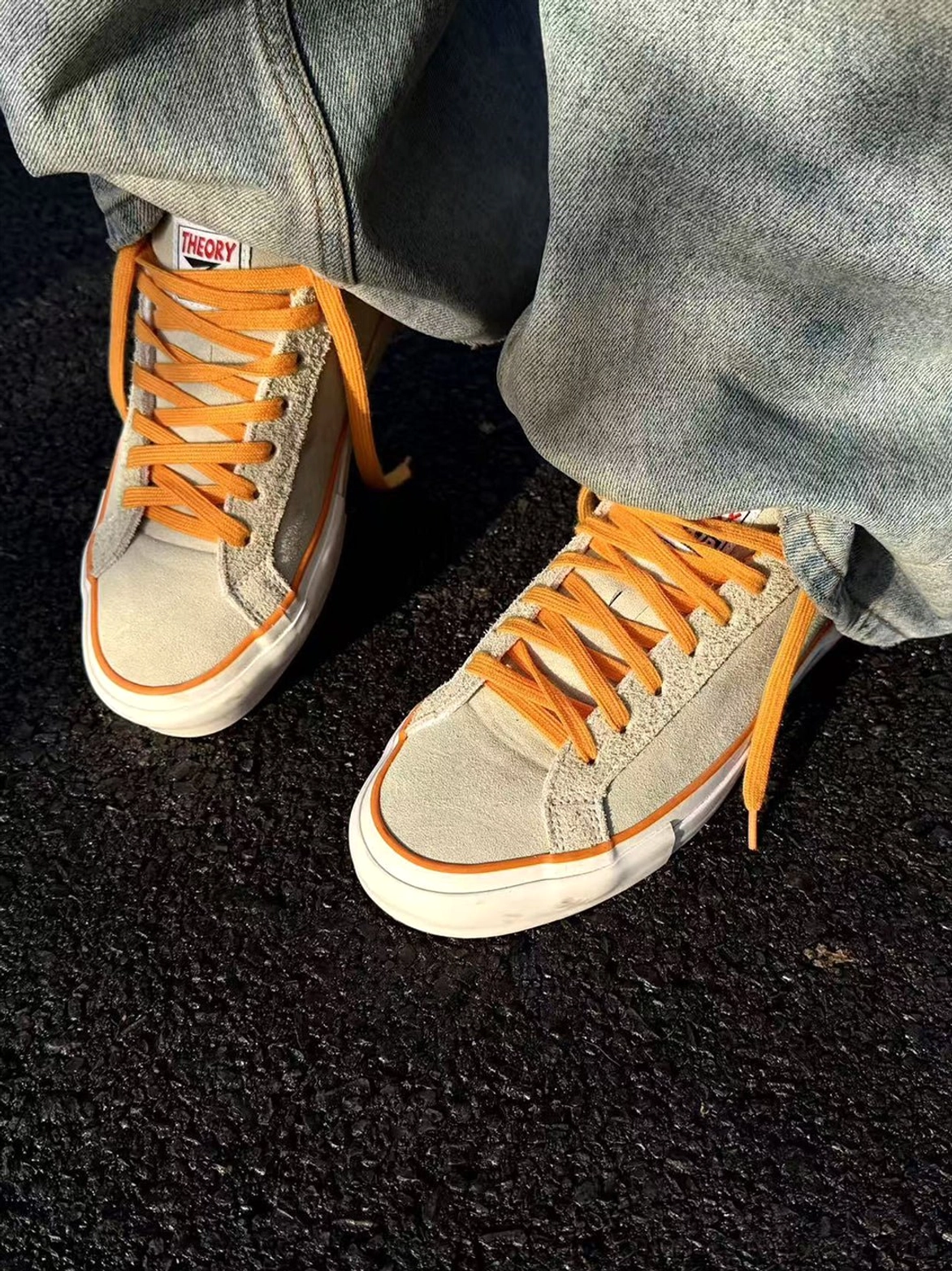 Close-up on-foot view of beige colorblock suede skate shoes with orange laces styled with wide-leg blue jeans