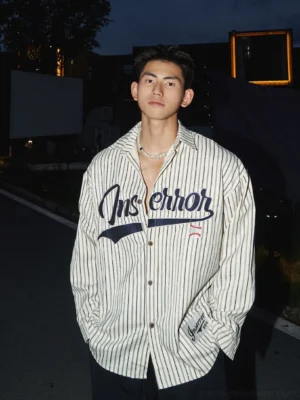 Beige oversized pinstripe baseball shirt worn with black trousers in a nighttime streetwear setting, featuring a button-down collar, vertical stripes, and embroidered sports-inspired chest graphic