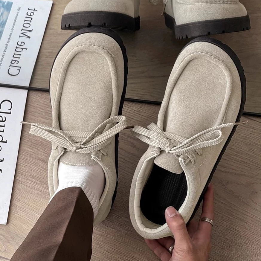 Beige square-toe suede lace-up streetwear shoes with matching laces, black ridged sole, white socks, brown trousers, and art magazine styling