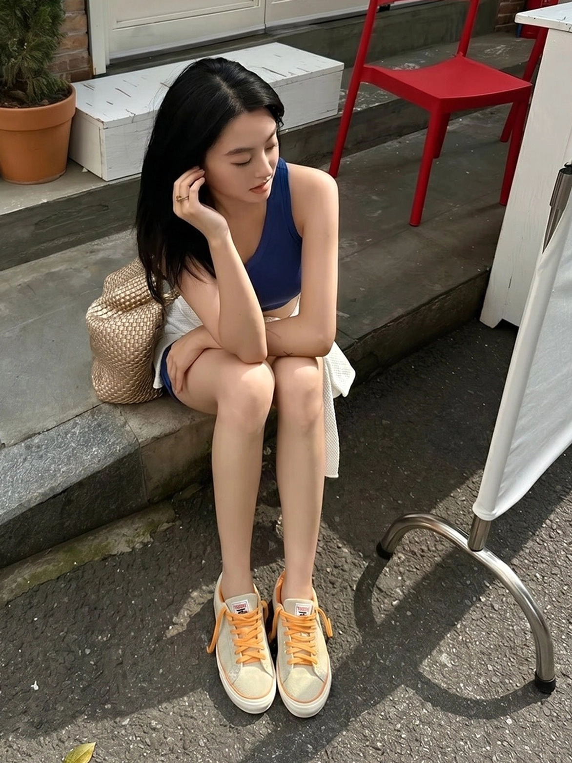 Female model wearing beige suede skate shoes with orange laces, white shorts, and a blue cropped top on outdoor cafe steps