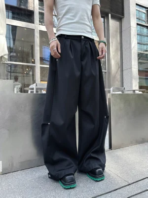 Front full-body view of black snap-pleat wide leg trousers styled on a male model in an urban street plaza