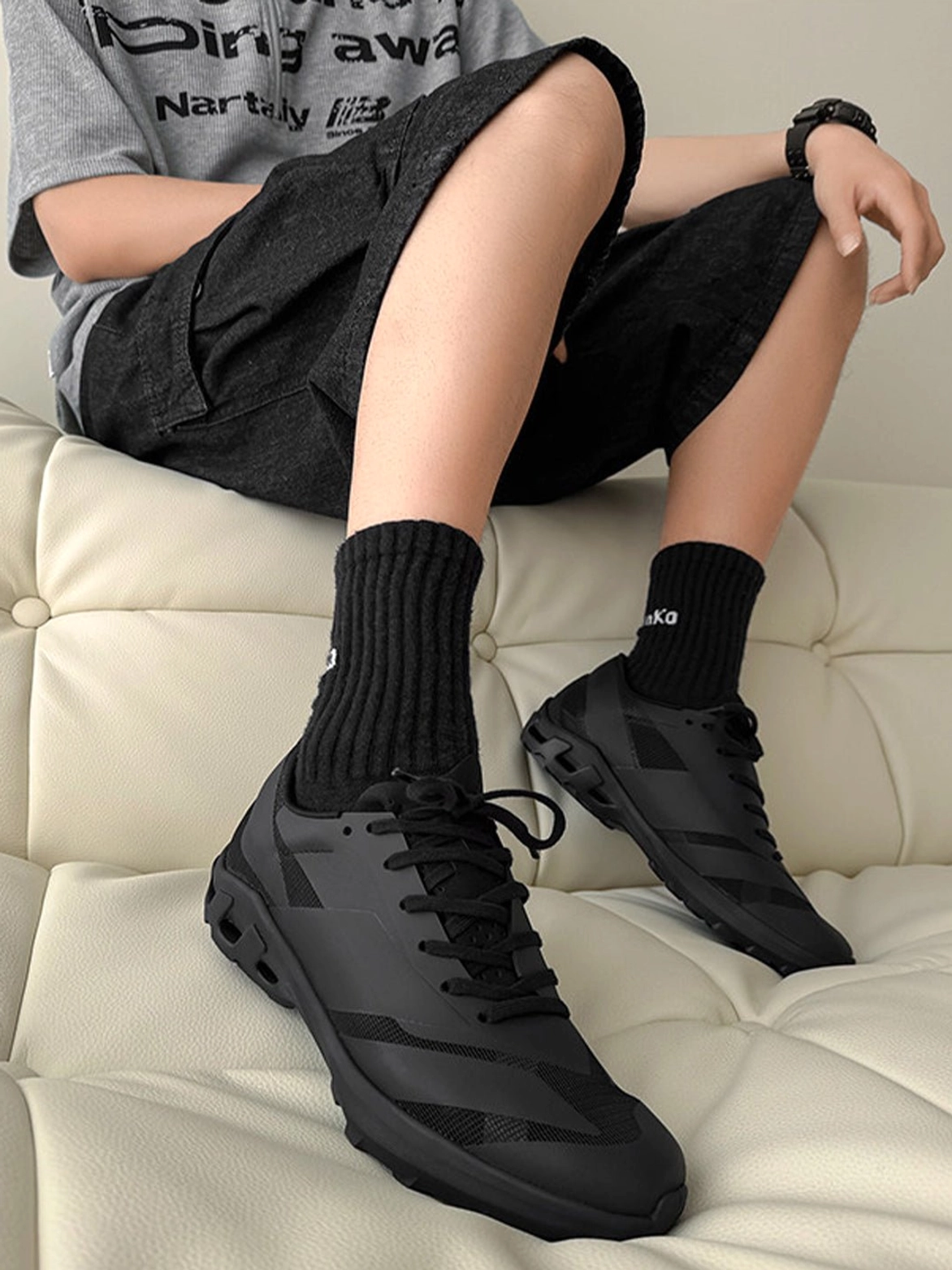 Male model wearing black low-profile arch-support sneakers with black socks, shorts, and a casual gray t-shirt in a relaxed streetwear outfit