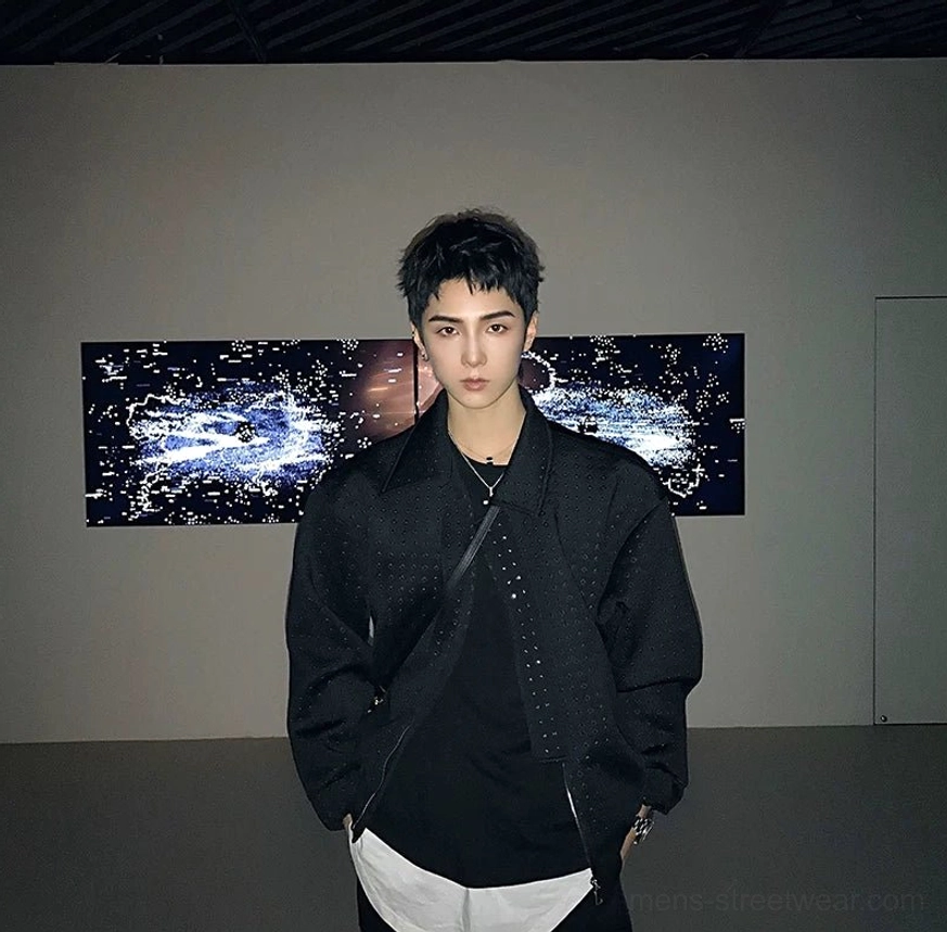 Black asymmetric zip techwear jacket front view styled with a crossbody chest strap and layered white hem in an art gallery