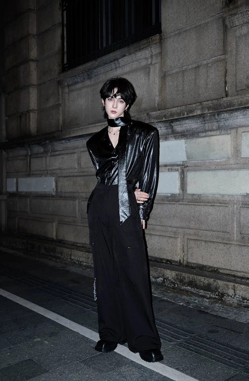 Androgynous model in an all-black avant-garde streetwear outfit with black boots, patent blazer, and wide-leg trousers against an urban stone wall