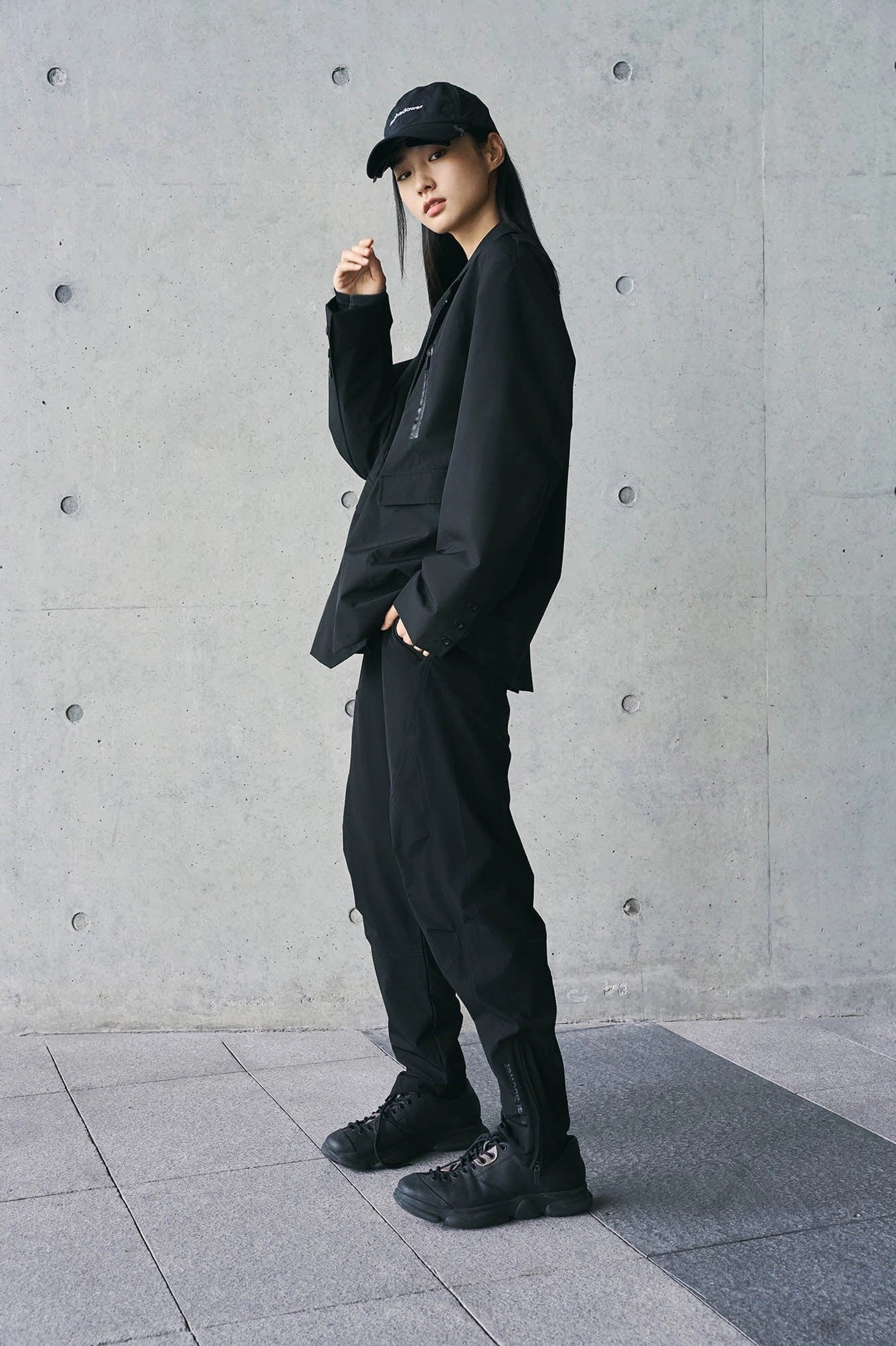 Three-quarter view of black avant-garde streetwear track pants styled with oversized technical jacket and cap