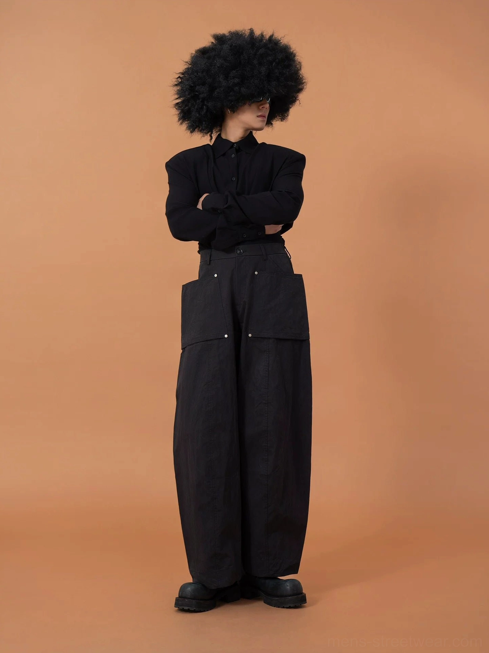 Model wearing black oversized cargo pod pants with oversized front pockets, metal studs, and a baggy balloon-leg streetwear silhouette