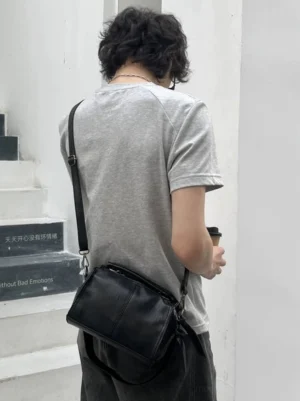 Compact black barrel shoulder bag with adjustable strap worn by male model in white indoor stairwell