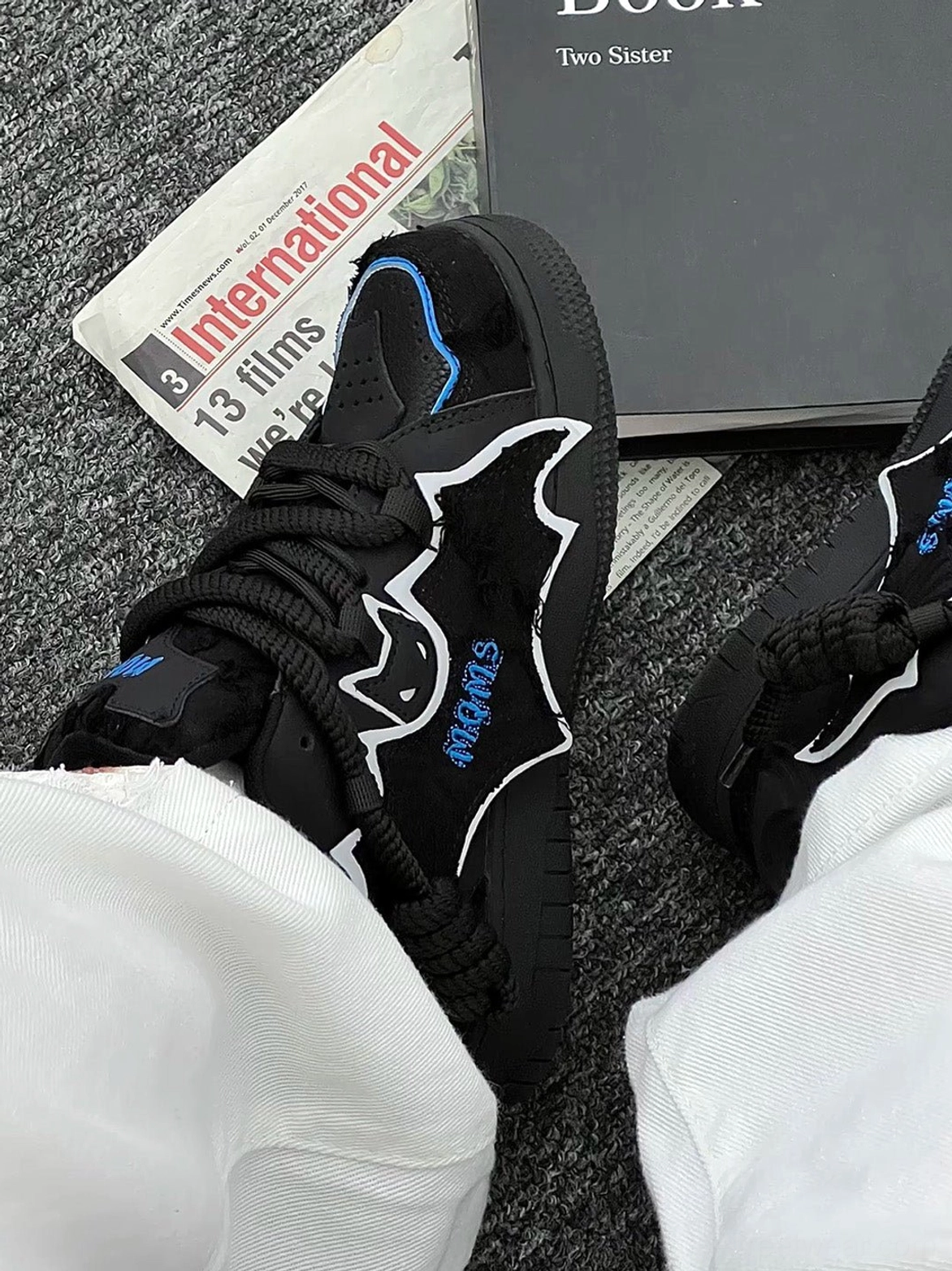 Black bat graphic platform sneaker shown from above with blue embroidery, fuzzy split leather texture, thick woven laces, white trousers, books, and fashion magazines