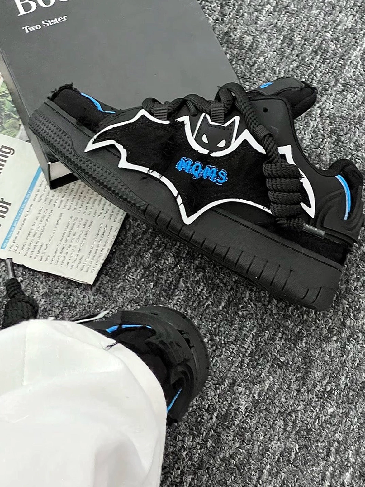 Black bat platform sneakers worn with white trousers and displayed on grey carpet, showing blue heel accents, outlined bat graphic, and thick rubber sole
