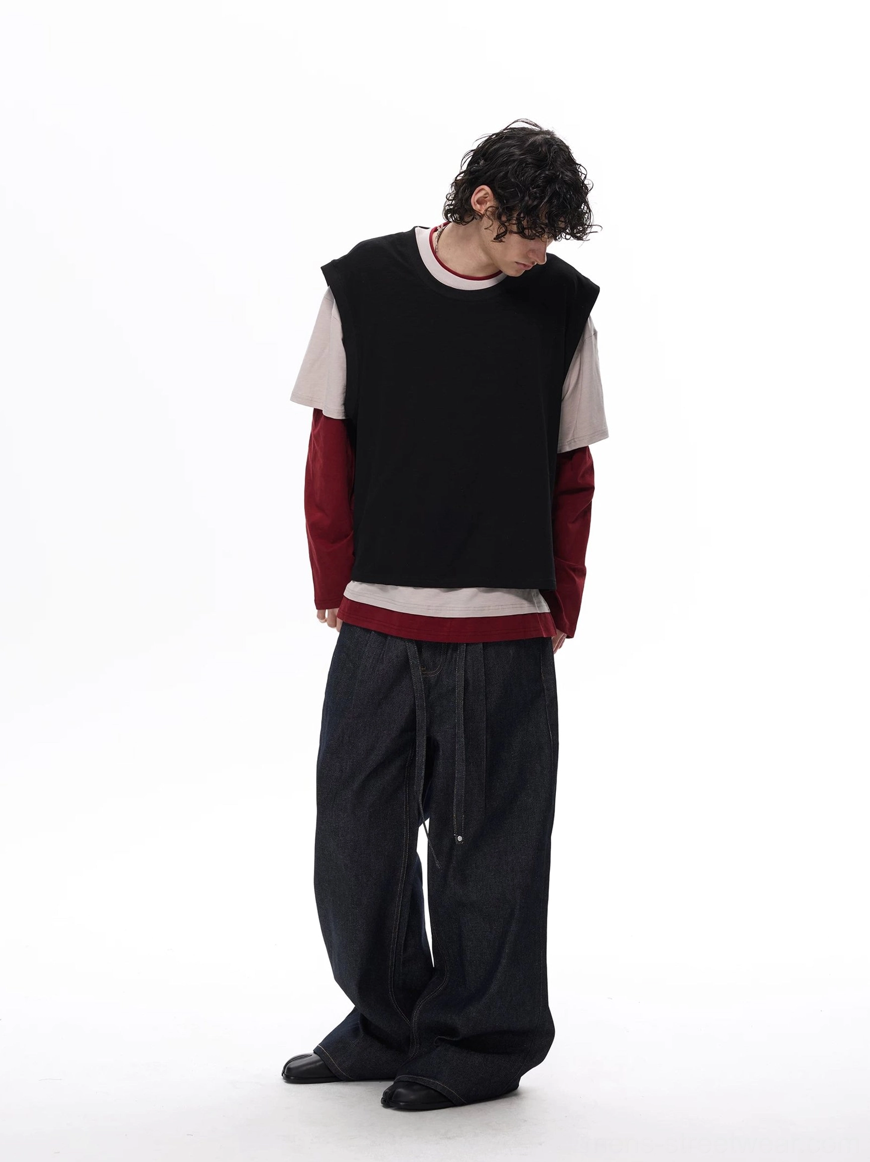 Right rear view of a male model in a black beige and burgundy layered vest tee top with relaxed dark denim streetwear styling
