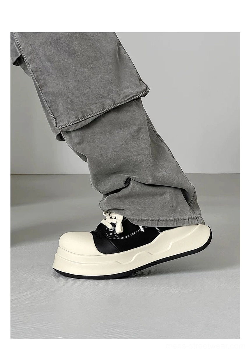 Side three-quarter wearing view of black big-head round toe sneakers with grey cargo trousers, curved toe cap, visible stitching, and layered platform sole