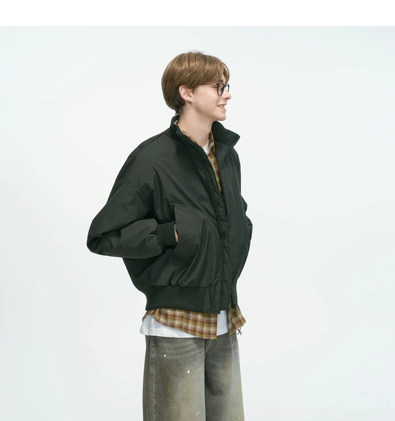 Black bomber jacket three-quarter side-back shot with hands in pockets, showing stand collar and ribbed trims, paired with plaid shirt and dirty-wash denim