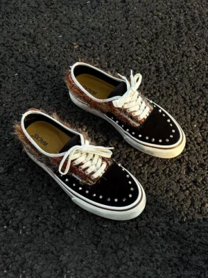 Black and brown mohair-panel studded low-top sneakers with fuzzy contrast panels, silver rivets, white laces, and rubber soles on an asphalt street