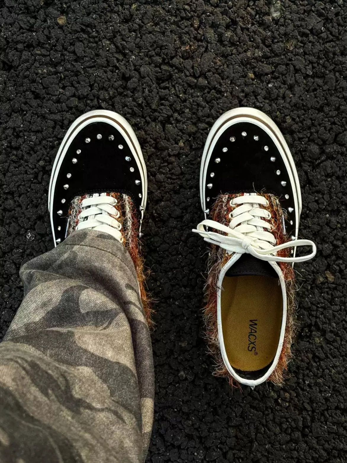Partial overhead view of black and brown studded low-top sneakers styled with camouflage cargo trousers and detailed textured toe caps