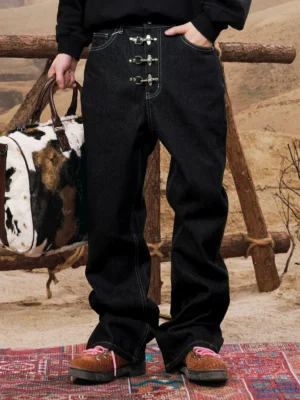 Front full-body view of black men's buckle wide leg jeans with contrast stitching and silver hardware styled in a desert streetwear setting