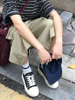 Black canvas platform sneakers with metal chain trim worn with white crew socks, striped shirt, and cropped khaki pants in an outdoor streetwear setting