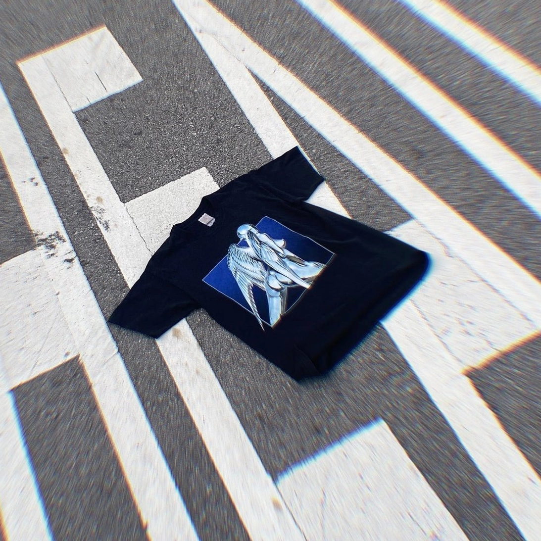 Angled flat lay of black chrome angel graphic t-shirt on city road markings with futuristic chromatic streetwear aesthetic