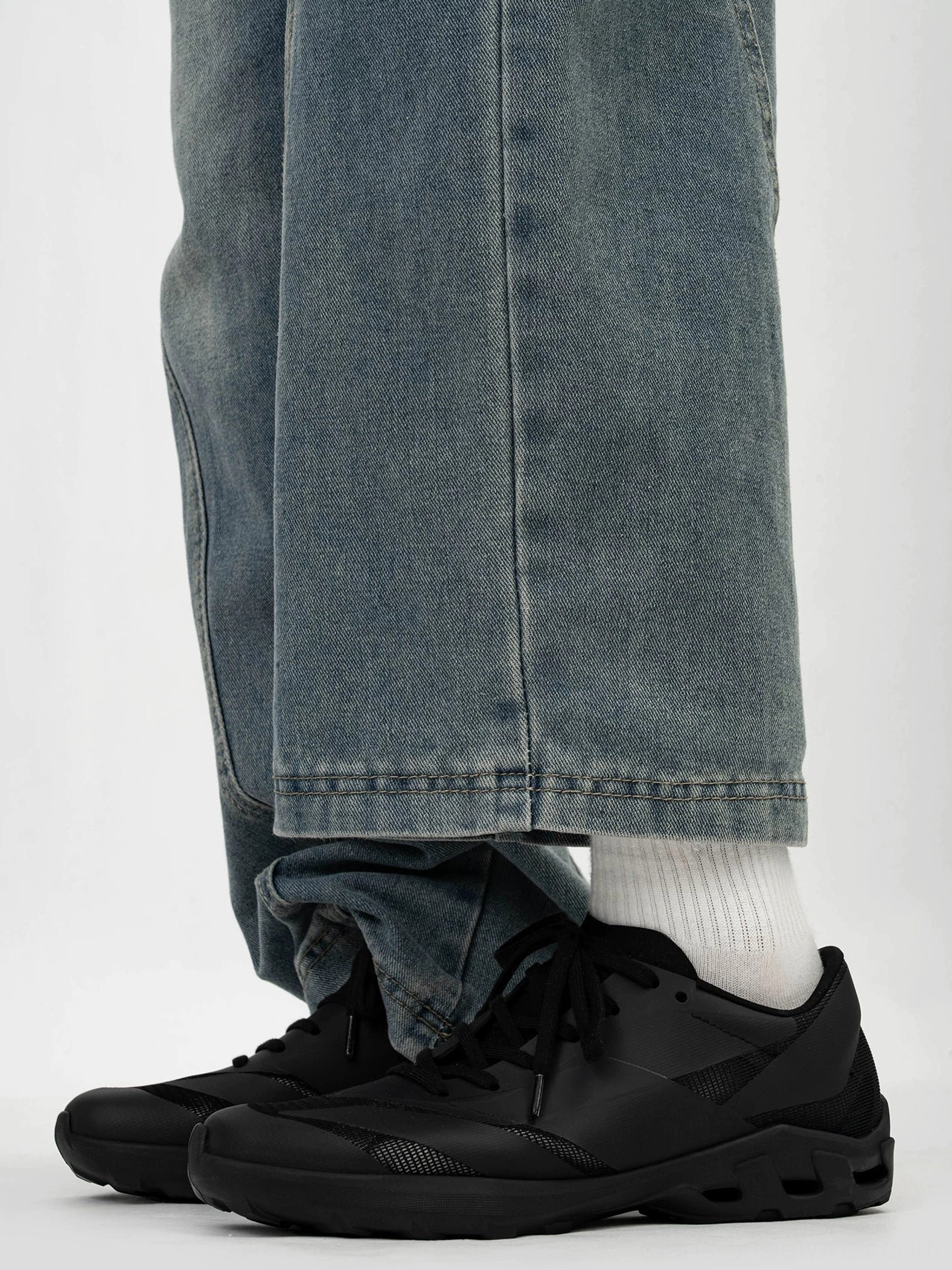 On-foot side view of black chunky trail sneakers with ventilated sole, matte seamless upper, cuffed blue jeans, and white crew socks