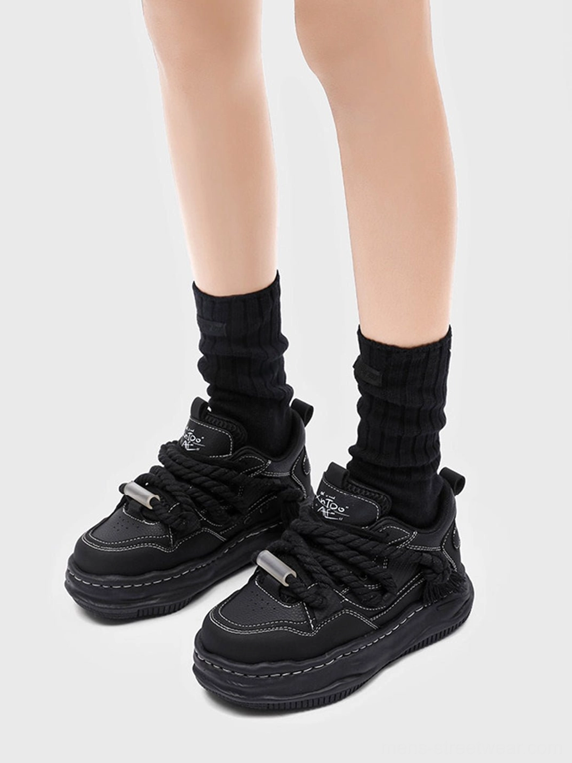 Front view of black chunky wavy platform sneakers worn with black crew socks, featuring twisted rope laces, metal toggles, and matte black upper