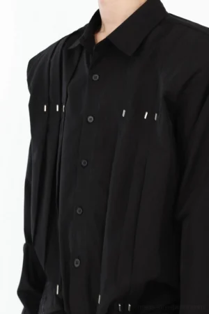 Close-up of black oversized cotton shirt showing classic collar, button closure, vertical pleats, silver metal rivets, and detailed hem