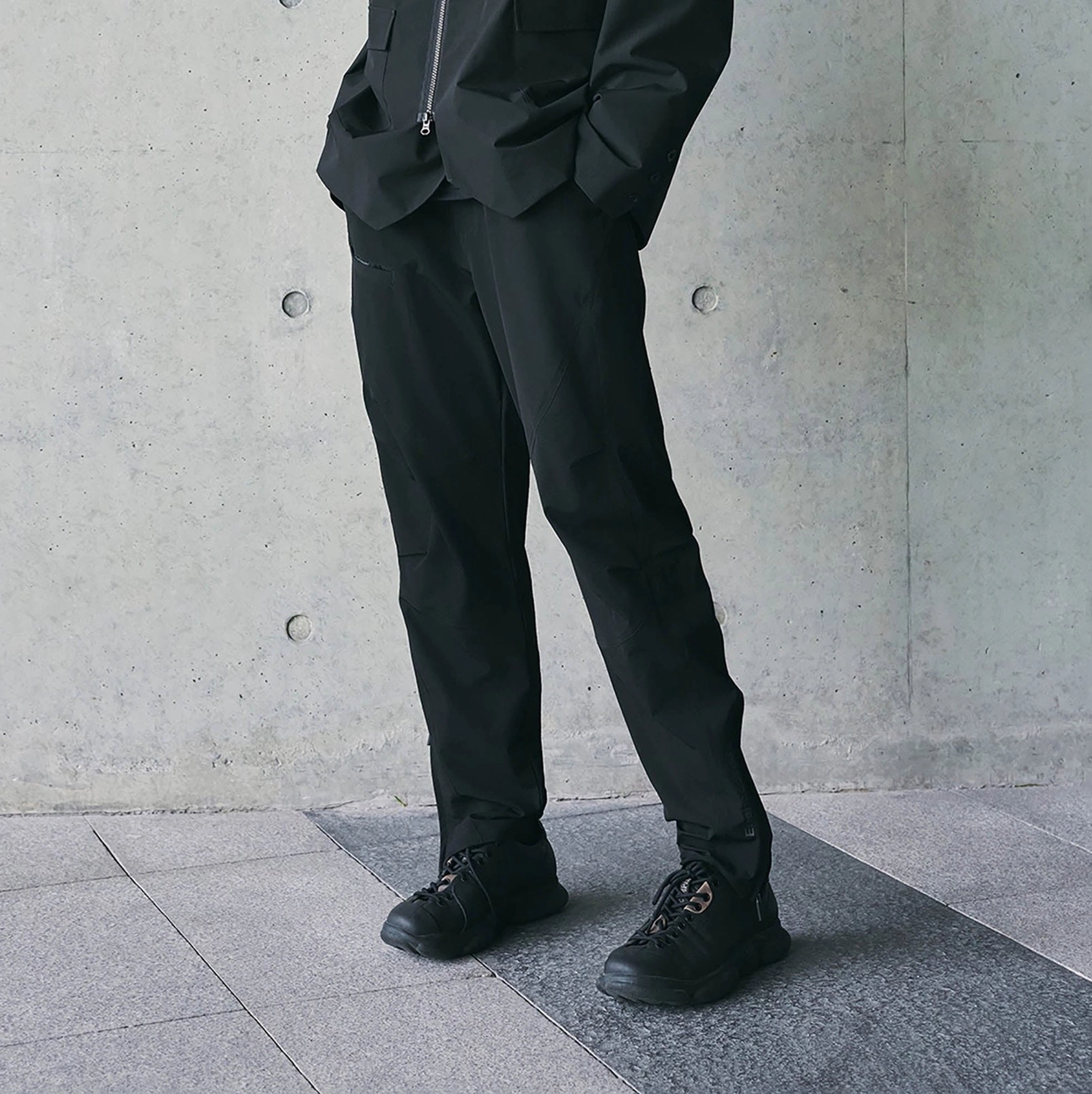 Side view of black crinkled techwear track pants featuring adjustable ankle zipper hems and matching technical jacket