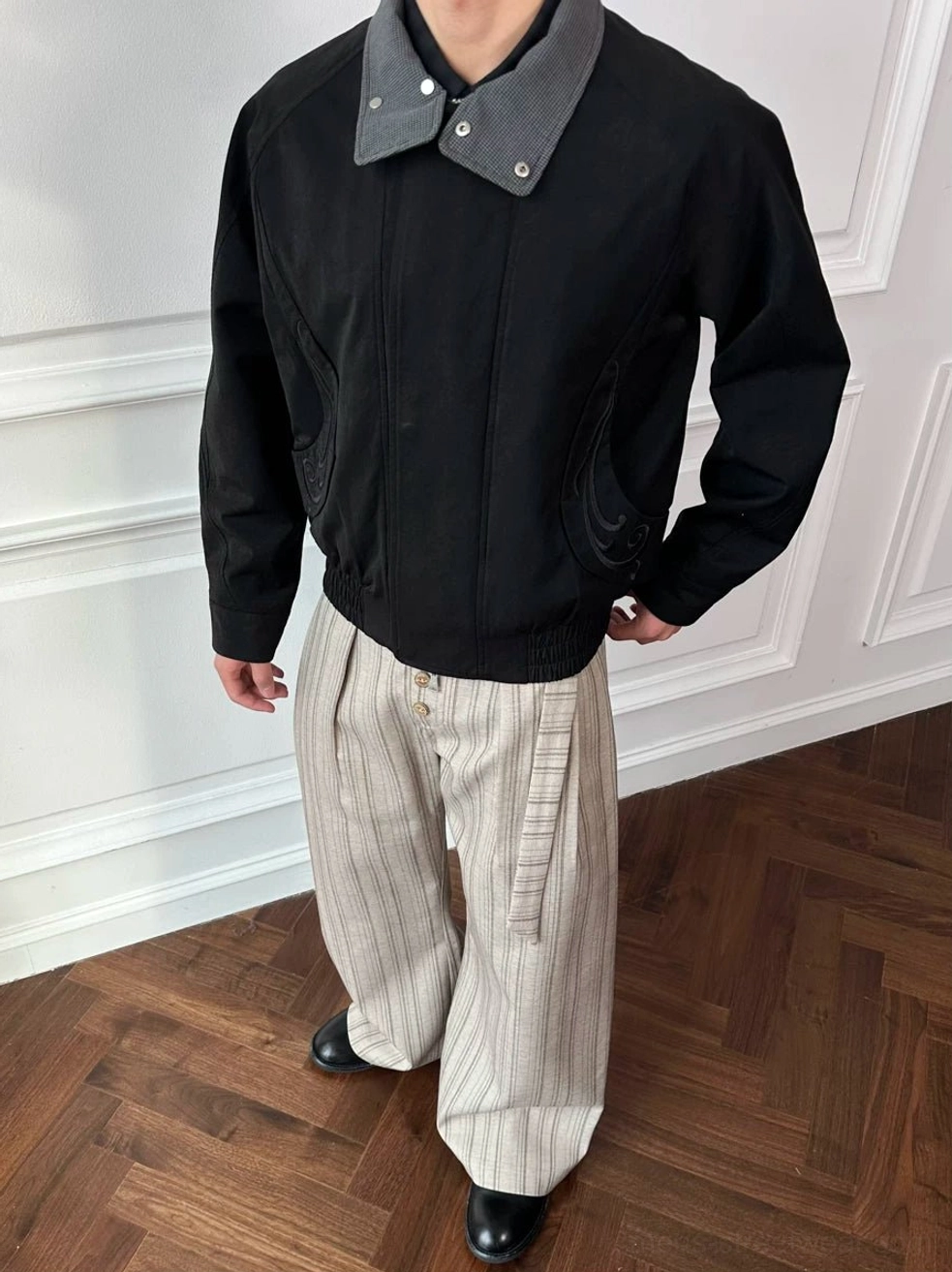 Male model wearing a black double-collar cropped bomber jacket with striped wide-leg pants and leather shoes in a vintage interior