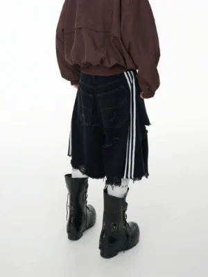 Rear three quarter view of black denim jorts with contrasting white side stripes, distressed detailing, frayed hems, brown cropped bomber jacket, and combat boots