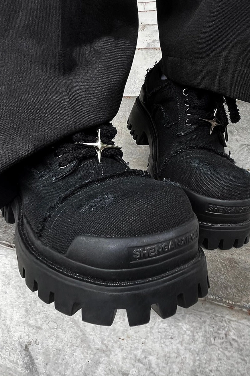 Low-angle close-up of black distressed canvas platform sneakers worn with wide-leg black trousers and chunky lug outsole