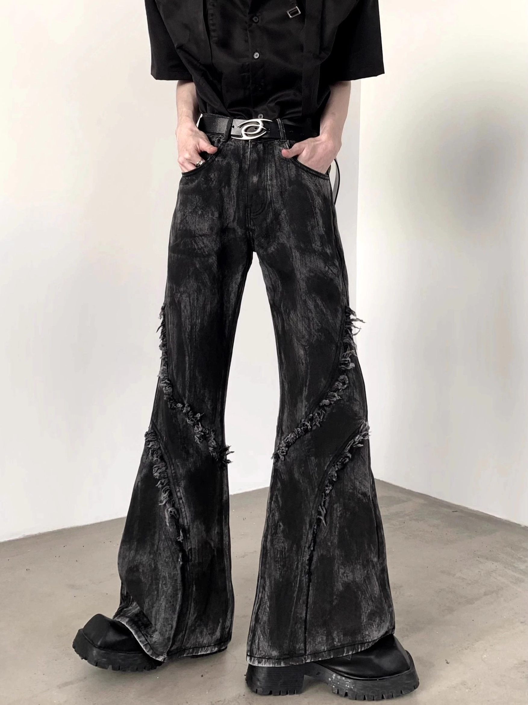 Front low angle view of black distressed flared jeans with faded denim wash, diagonal frayed seams, wide flare legs, black shirt, and platform boots