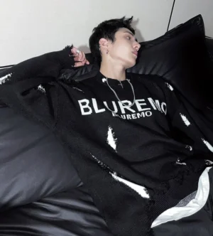 Side profile of a relaxed black distressed knit sweater showing oversized dropped shoulders, scattered shredded accents, and layered streetwear styling