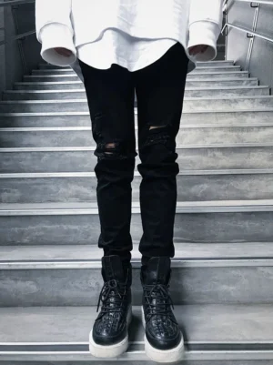 Front lower-body view of black distressed skinny jeans with frayed knee rips and tapered stacked hems worn with black boots on concrete stairs