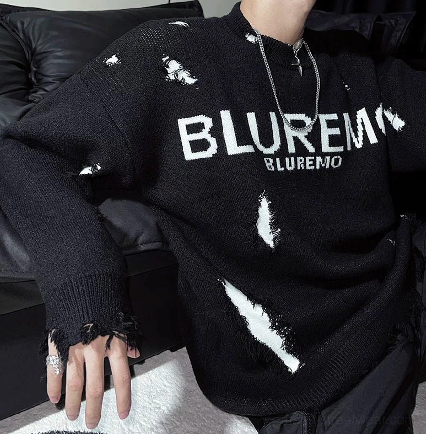 Close-up three-quarter view of a black distressed streetwear sweater showing frayed ribbed cuff, shredded knit cutouts, chest lettering, and layered necklaces