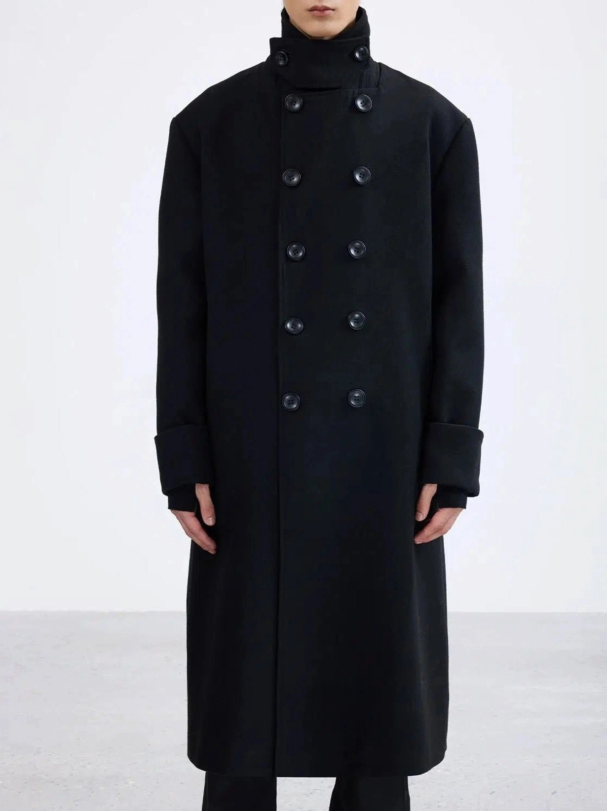 Black double-breasted long overcoat front close-up showing high collar button fastening wide cuffs and tailored streetwear construction