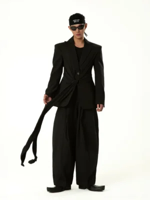 Front three-quarter view of a black draped tie-front blazer coat with structured shoulders, single-button closure, long waist sash, and avant-garde Korean streetwear styling