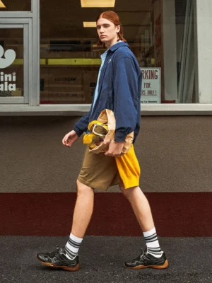 Male model wearing black drawcord runner sneakers with a navy jacket, light blue shirt, khaki shorts, and striped crew socks on an urban street