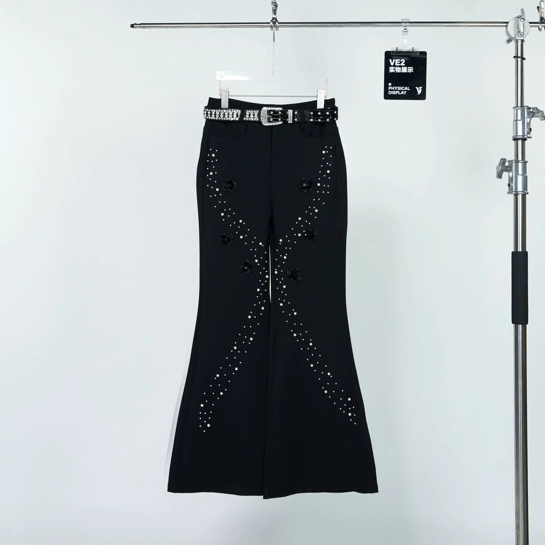 Black embellished flare trousers hanging on a clothing rack with matching studded belt, pearl-style star trails, and 3D floral applique details