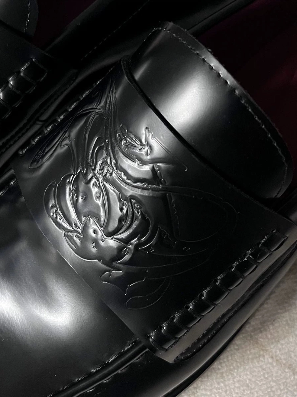 Close-up of glossy black embossed leather loafer vamp showing decorative texture, refined stitching, and contemporary square-toe styling