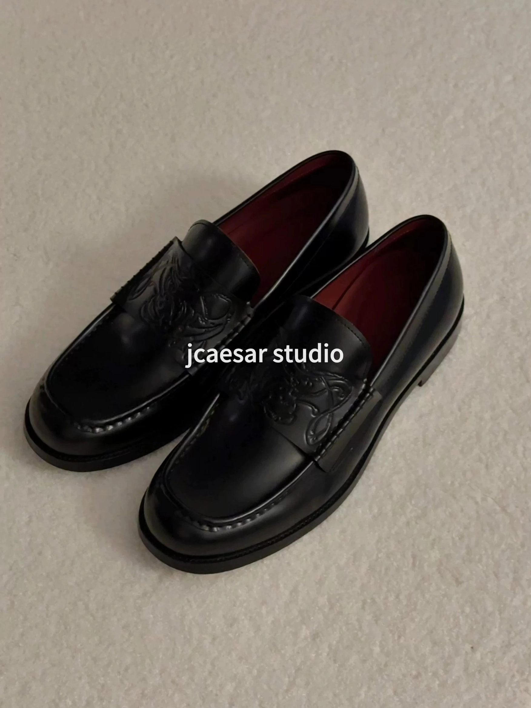 Black embossed leather loafers displayed on a light beige fabric surface with glossy uppers and contrasting burgundy lining